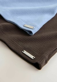 Tissu côtelé plié en bleu clair et marron foncé, chacun avec une petite étiquette métallique rectangulaire portant la mention « STUDIO » près du bord.