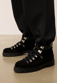 Juodi zomšiniai žygio batai su metaliniais aklais ir storomis guminėmis padangomis, avimi su juodomis kelnėmis ant lygaus smėlio spalvos grindų.