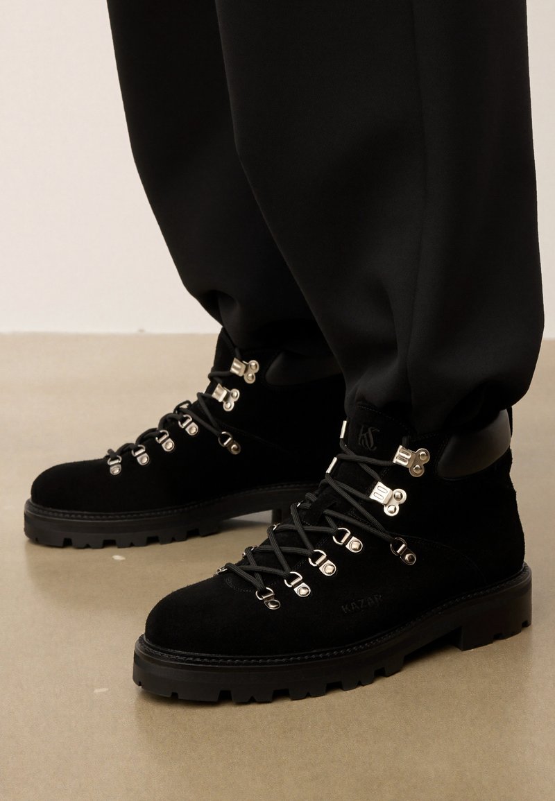 Juodi zomšiniai žygio batai su metaliniais aklais ir storomis guminėmis padangomis, avimi su juodomis kelnėmis ant lygaus smėlio spalvos grindų.