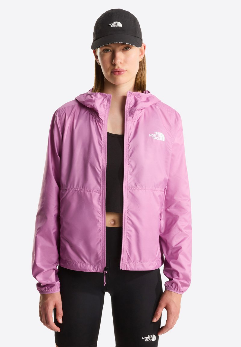Nainen pukeutuneena vaaleanpunaiseen hupulliseen takkiin, mustaan urheiluliiviin, mustiin legginsseihin ja mustaan lippalakkiin, jossa on The North Face -logot, seisomassa valkoista taustaa vasten.