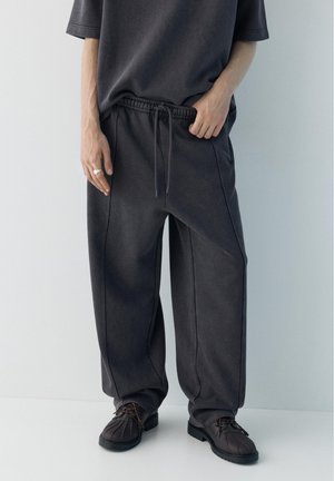 Persona che indossa pantaloni della tuta larghi grigio scuro con coulisse e scarpe nere con lacci, in piedi davanti a uno sfondo chiaro uniforme.