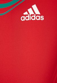 Camiseta atlética roja de tela suave, que presenta una franja de acento verde y el logo blanco de Adidas en el frente.