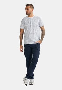 Hvid kortærmet t-shirt med bladmønster, båret med mørkeblå cargo bukser og hvide sportsko. Har rund hals og et subtilt logo.