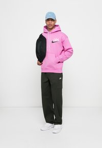 Felpa rosa con logo, borsa a tracolla nera, pantaloni cargo verdi oliva e sneakers bianche. Un cappellino blu completa l'outfit.