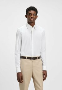 Chemise blanche à manches longues avec un col pointu, associée à un pantalon beige ajusté et une ceinture marron avec une boucle en argent.