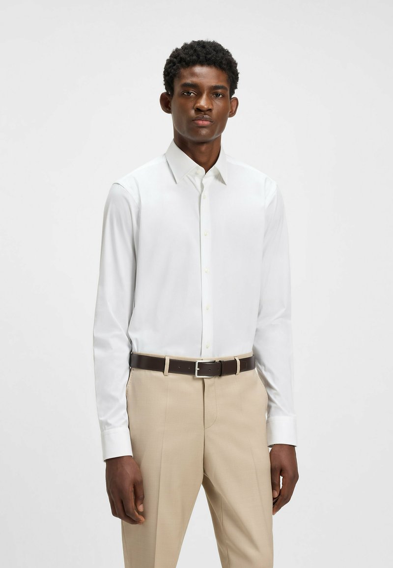 Chemise blanche à manches longues avec un col pointu, associée à un pantalon beige ajusté et une ceinture marron avec une boucle en argent.