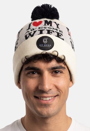 Gebreide muts in roomkleur met rode en zwarte patronen, met de tekst "IK HOU VAN MIJN GEWELDIGE VROUW" en een zwart logo-patch. Bovenop zit een zwarte pompon.