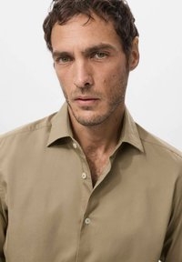 Camisa de botones color beige con cuello, que muestra una textura suave y una tela blanda. Dos botones visibles en la tapeta, corte moderno.