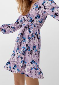 Robe fleurie en lilas avec des accents bleus et noirs. Caractéristiques : manches longues, col en V, détail de ceinture et volants superposés à l'ourlet.