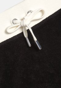 Κοντινό πλάνο μαύρου υφάσματος με λευκή ζώνη στη μέση που έχει δεμένο λευκό κορδόνι με μεταλλικά άκρα μέσα από ασημένια αυτιά.