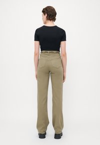 Person med kort krøllet hår iført en tettsittende svart t-skjorte gjemt i høyhalsede olivengrønne jeans og svarte platå sko, stående vendt bort.