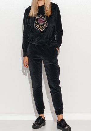 Kobieta ubrana w czarny aksamitny dres z emblematem róży i napisem "MAKADAMIA", w połączeniu z czarnymi, ćwiekowanymi trampkami, stoi na tle białej ściany.