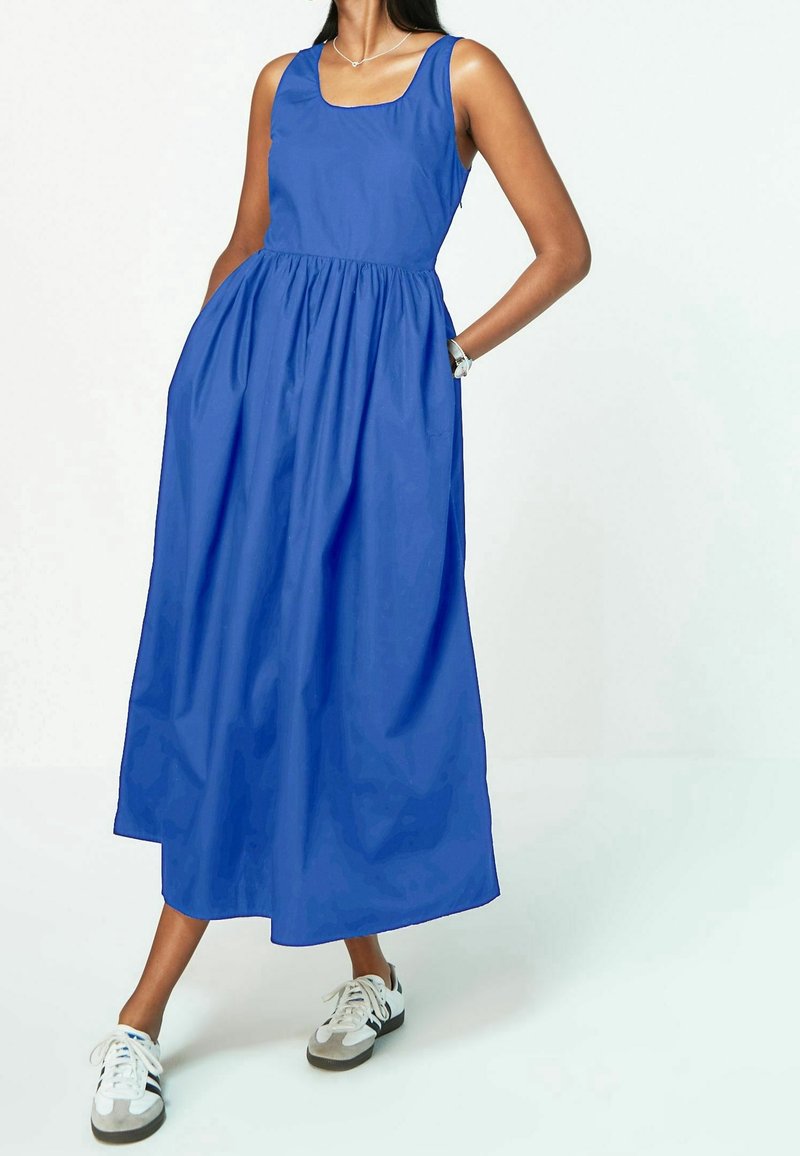 Blue sleeveless dress with a square neckline, gathered waist, and full skirt. Worn with grey and white athletic shoes.