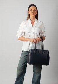 Sac à main en cuir noir avec une texture lisse, une forme arrondie et deux poignées robustes. Présente un accent logo doré à l'avant.
