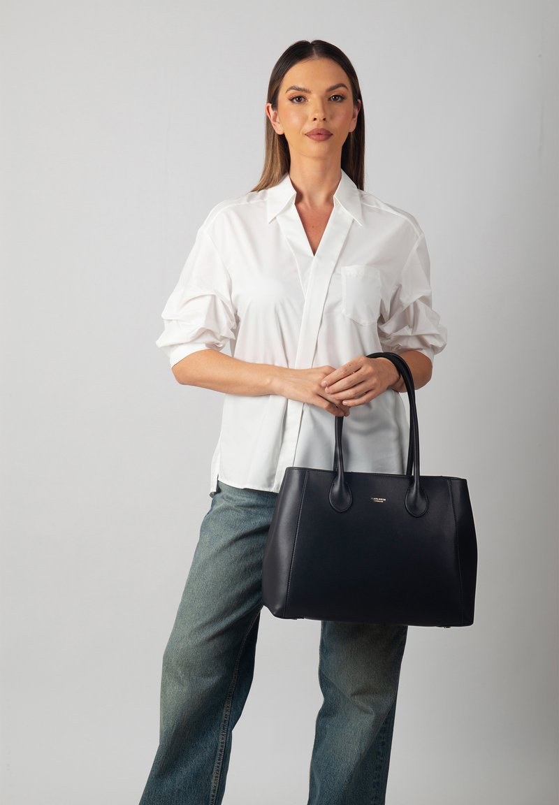 Sac à main en cuir noir avec une texture lisse, une forme arrondie et deux poignées robustes. Présente un accent logo doré à l'avant.