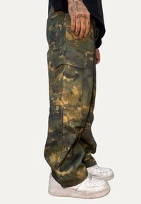 Pantaloni cargo con motivo mimetico verde, realizzati in tessuto pesante, dotati di multiple tasche e vestibilità rilassata, abbinati a sneakers bianche.