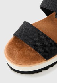 Sandal din pânză neagră cu brâu, cu o suprafață din suede maro și o talpă intermediară groasă albă. Detalii texturate din material și cauciuc pe talpă.