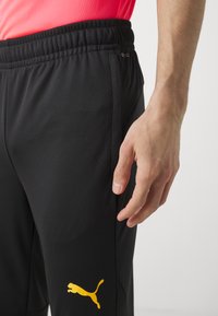Zwarte sportbroek met een gestructureerde afwerking en een geel Puma-logo op het linkerdij, voorzien van een elastische tailleband voor comfort.