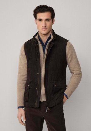 Gequiltete dunkelbraune Weste über einem beigen Strickpullover, kombiniert mit einem blau karierten Hemd. Verfügt über zwei Fronttaschen und einen Reißverschluss.