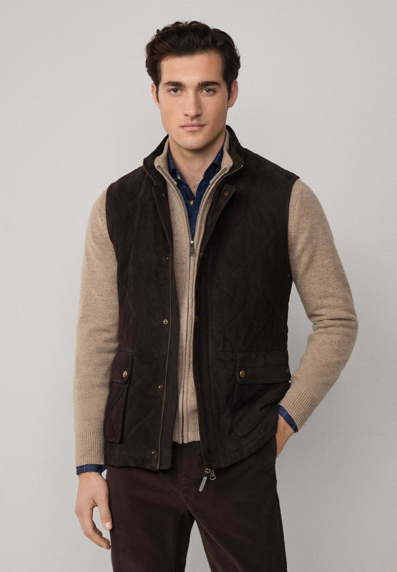 Gilet trapuntato marrone scuro sopra un maglione beige a maglia, abbinato a una camicia a quadri blu. Presenta due tasche anteriori e una chiusura zip.