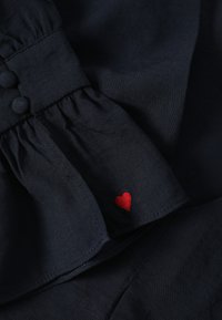 Tissu bleu marine foncé avec poignet froncé et boutons recouverts de tissu, présentant un petit cœur rouge brodé près du bord.