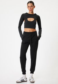 Schwarzes, gekürztes Langarmoberteil mit Ausschnittdesign, kombiniert mit schwarzen Jogginghosen mit elastischem Bund und seitlichen Taschen, getragen mit Turnschuhen.