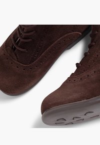 Zapatos de ante marrón oscuro con diseño de cordones, con detalles perforados a lo largo de los costados y una suela de goma con un fondo texturizado.