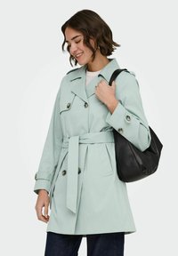 Femme en trench-coat bleu clair ceinturé tenant un sac à bandoulière noir, souriante et regardant vers le bas, portant un pantalon foncé et un haut blanc.