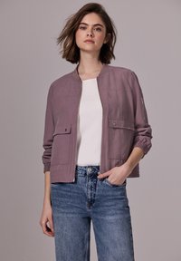 Lila bomberjacka med dragkedja framtill, elastiska muddarna, två framfickor och en mjuk textur, bärs över en vit skjorta och blå jeans.