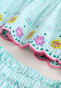 Vitt tyg med ljus turkosa ränder och färgglada broderade blommor, med rosa vågiga kanter och veckad elastisk midjeresår.