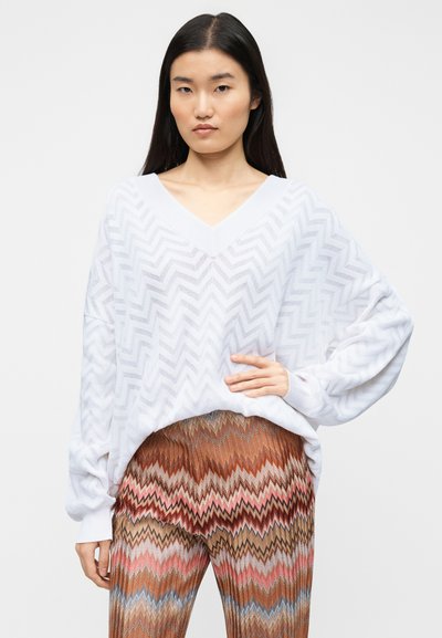 Λευκό πλεκτό πουλόβερ με V λαιμόκοψη και σχέδιο σε σχήμα κεραυνό. Συνδυασμένο με καφέ παντελόνι με ζιγκ ζαγκ μοτίβο και κόκκινες και μπλε λεπτομέρειες.