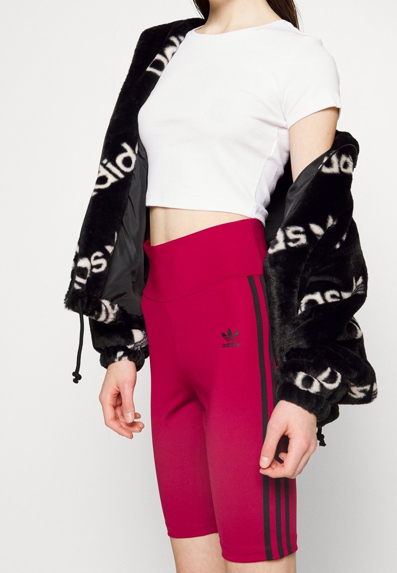 Černá bunda z umělé kožešiny s bílou podšívkou s logem, bílý krátký top a růžové cyklistické šortky s vysokým pasem s černými bočními pruhy a logem.