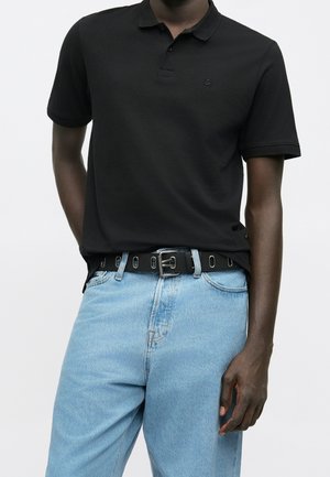 Mężczyzna w czarnej koszulce polo z krótkim rękawem, wpuszczonej w jasnoniebieskie dżinsy z czarnym paskiem, stojący na tle jasnego, jednolitego tła.