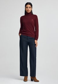 Eine Frau steht da und trägt einen burgunderfarbenen Rollkragenpullover, navyfarbene, weite Hosen und braune, knöchelhohe Stiefel mit Absatz vor einem einfarbigen Hintergrund.