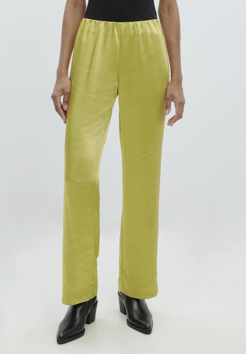 Limoengroene elastische broek met rechte pijpen en een gladde textuur, gedragen met zwarte hakken, weergegeven tegen een lichte achtergrond.