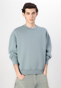 Fleece crewneck sweatshirt in gedempt teal. Heeft ribgebreide manchetten en zoom, met subtiele branding op de borst. Zachte textuur en een ontspannen pasvorm.