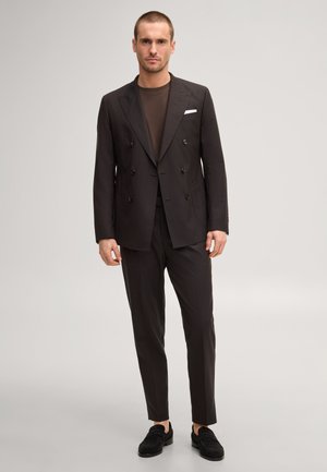 Mann trägt einen dunklen, zweireihigen Anzug mit braunem Hemd und schwarzen Loafers und steht vor einem neutralen Hintergrund.