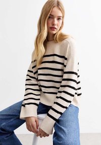 Maglione beige con strisce orizzontali nere, collo a girocolo rigato e maniche oversize, abbinato a jeans di denim blu. Morbido tessuto a maglia.