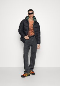 Svart dunjakke med grønn hetteforing, oransje grafisk t-skjorte, mørke jeans og livlige flerfargede joggesko. Ingen synlige merkevarer.