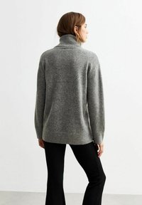 Grijze gebreide trui met een hoge kraag, ribgebreide manchetten en een iets langere achterkant, gecombineerd met zwarte broek.