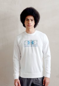 Sudadera blanca de manga larga con logo azul y el texto "UNDER ARMOUR" en la parte frontal, hecha de una tela suave y lisa, con puños y dobladillo acanalados.