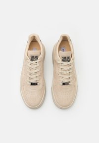 Pár béžových semišových tenisek s perforovanými špičkami a béžovými tkaničkami, pohled zhora na bílém pozadí.