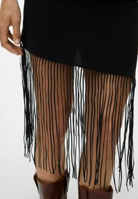 Jupe noire avec de longues franges au bas, mettant en valeur une texture de tissu lisse. Main positionnée sur la frange, bottes en cuir marron en dessous.