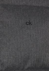 Sort tekstil med fiskebenmønster og diskret glans. Har "ck"-logo, glat tekstur og struktureret form. Ideel til dekorativ brug.