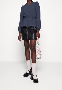 Femme portant un chemisier bleu marine à manches longues, une mini-jupe en cuir noire, des chaussettes blanches, des chaussures Mary Jane noires à talons, tenant un petit sac blanc avec un imprimé orange.
