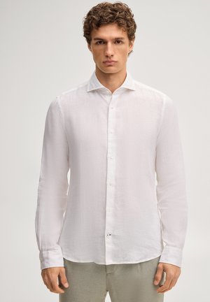 Giovane uomo con capelli ricci che indossa una camicia bianca con bottoni e maniche lunghe e pantaloni grigio chiaro, in piedi davanti a uno sfondo semplice.