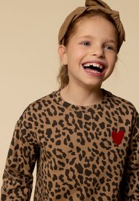 Lachend jong meisje met een bruine haarband en een top met luipaardprint en een rode hart op de borst tegen een neutrale achtergrond.