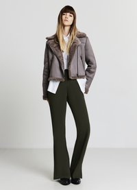 Giacca grigia in pelle sintetica con colletto in montone marrone, camicia bianca sotto e pantaloni verdi scuro a gamba larga, abbinati a scarpe nere.