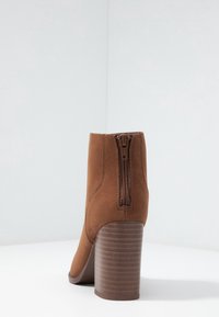 Brązowy zamszowy botek za kostkę z tylnym zamkiem błyskawicznym i masywnym, żeberkowanym wysokim obcasem. Elegancki design z gładkim wykończeniem i zaokrąglonym noskiem.