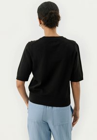 Schwarzer Kurzarmpullover mit geripptem Ausschnitt und Saum, von hinten gezeigt, kombiniert mit hellblauen Hosen. Glatter Strickstoff.